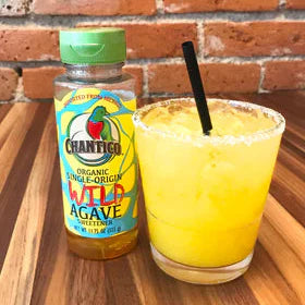 Bottle of Chantico agave nectar next to a glass of yellow drink with a straw on a wooden surface.