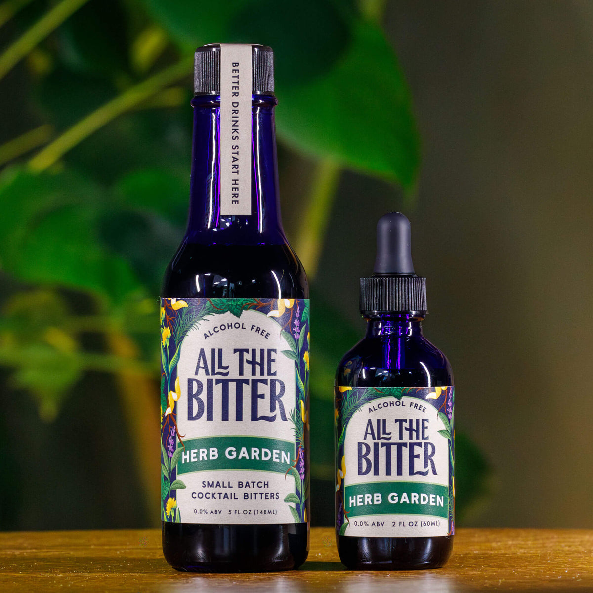 Two bottles of 'All The Bitter Herb Garden' bitters on a wooden surface with a blurred green background.